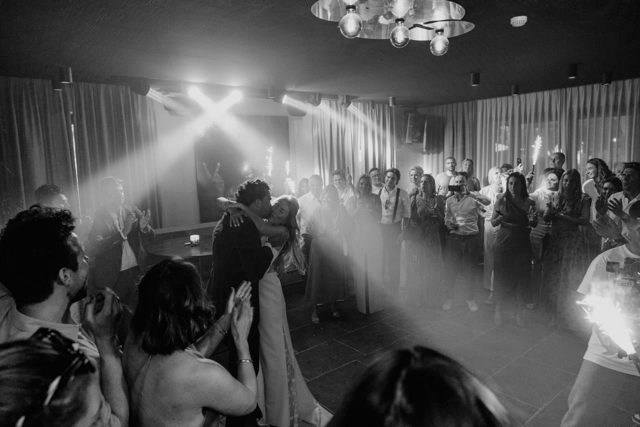 First dance: the couple kissing in a beam of light, surrounded by guests with sparklers, in black and white